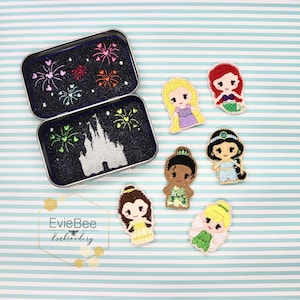 Op de afbeelding: Een set van zeven geborduurde vingerpoppen met Disney-prinsessen. De prinsessen zijn Ariel, Belle, Assepoester, Jasmine, Mulan, Rapunzel en Tiana. De poppen zitten in een klein blikje met een zwarte achtergrond en een wit silhouet van Assepoesters kasteel met vuurwerk.