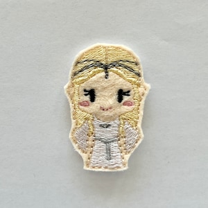 May include: Embroidered patch of a cartoon girl with blonde hair, wearing a white dress and a crown. She has black eyes and pink cheeks.
