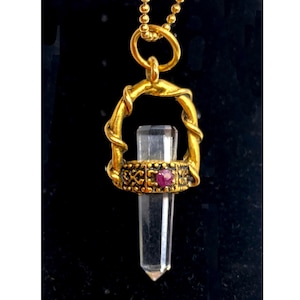 May include: A gold-colored pendant necklace featuring a clear crystal point. The crystal is held within a decorative gold setting with a red gemstone accent. The necklace hangs from a delicate gold chain against a black background.