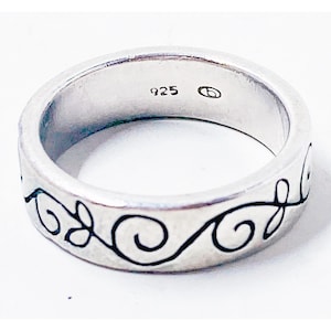 Può includere: Un anello d'argento con un motivo floreale nero. L'anello è contrassegnato con "925". La fascia presenta un motivo ripetuto di foglie e volute. L'anello è uno stile a fascia semplice.