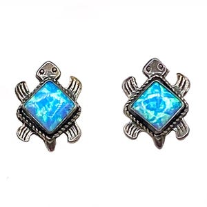 Könnte beinhalten: Ein Paar silberfarbene Turtle-Ohrstecker. Jeder Ohrring hat einen quadratischen, hellblauen Opalstein, der in einen schildkrötenförmigen Rahmen gefasst ist. Das Schildkrötendesign umfasst einen Kopf, Flossen und einen seilartigen Rand.