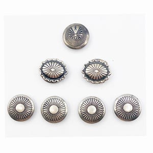 May include: A collection of silver-toned metal buttons with intricate designs. The buttons feature various patterns, including a central sunburst design and a spiral motif. The buttons are arranged on a white background, showcasing their detailed craftsmanship.