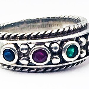 May include: Silver ring with a braided rope design and a row of colorful gemstones. The ring features a dark blue, red, green, and purple gemstone. The ring is a piece of jewelry.