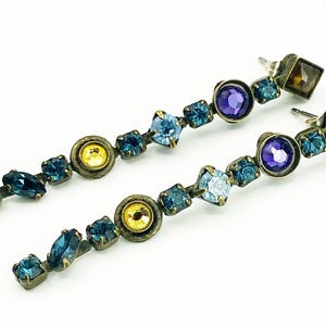 May include: A pair of drop earrings with a linear design. The earrings feature blue, purple, and yellow gemstones. The metal has an antique brass finish. The earrings are displayed against a white background.