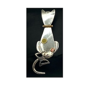 May include: A silver cat-shaped brooch with gold accents. The brooch features a stylized cat design with a gold collar, a gold circle, and a red and gold accent. The tail is a curved silver shape with a bow.