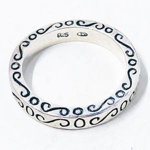 Può includere: Un anello d'argento con un motivo ripetuto di volute e cerchi neri. L'anello è contrassegnato con "925" e un piccolo simbolo. L'anello è uno stile a fascia, con il design che avvolge l'intera circonferenza.