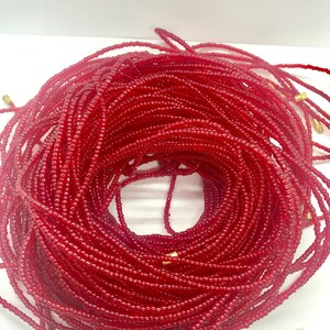 May include: A close-up of a red beaded waist beads, also known as waist beads or belly beads. The beads are strung on a thin cord and are tied together with a gold-colored clasp. The beads are arranged in a circular shape, creating a beautiful and intricate design.