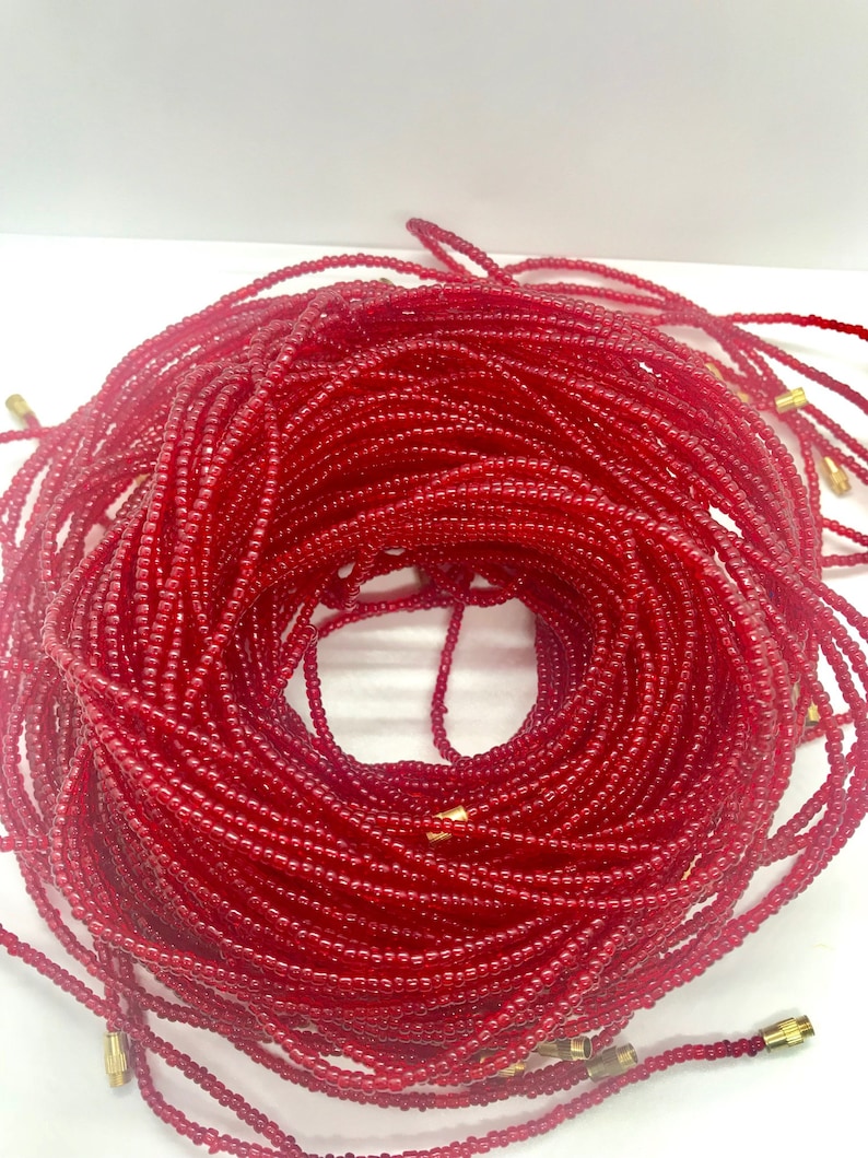 May include: A close-up of a red beaded waist beads, also known as waist beads or belly beads. The beads are strung on a thin cord and are tied together with a gold-colored clasp. The beads are arranged in a circular shape, creating a beautiful and intricate design.