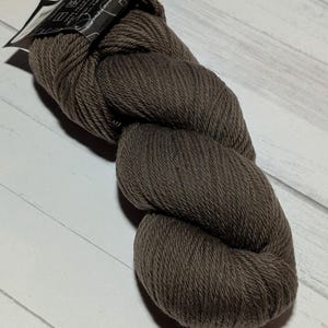 Op de afbeelding: Een streng donkerbruin garen gemaakt van 100% Peruaanse hooglandwol. Het garen is gedraaid tot een dikke, getextureerde kluwen. Aan de bovenkant zit een zwart papieren label met witte tekst, waaronder de merknaam Cascade.