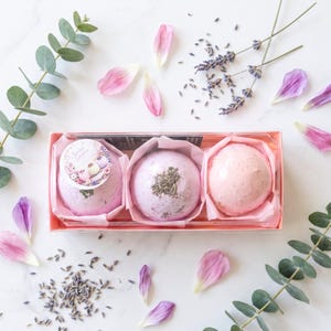 May include: A rectangular box containing three round bath bombs in shades of pink and lavender, decorated with dried lavender and flower petals. The text reads: Transform your stress into a luxurious floral escape. The perfect ready-to-give gift. Treat Yourself.
