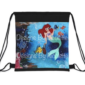 May include: Black drawstring bag with a colorful illustration of Ariel the Little Mermaid swimming with Flounder and other fish. The bag has the text "Designs By Nicolette" printed on it.