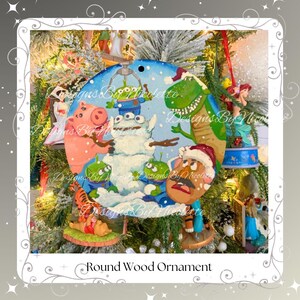 May include: Round wood ornament featuring a winter scene with Toy Story characters. The ornament has a blue sky background and a snowman with the aliens, Rex, Mr. Potato Head, and Hamm. The ornament is hanging on a Christmas tree.