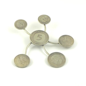 Könnte beinhalten: Ein silberfarbener Münzhalter mit einer zentralen Münze mit der Zahl "5" und der Aufschrift "Bundesrepublik Deutschland". Fünf weitere Münzen sind angebracht, jede mit unterschiedlichen Designs und Werten. Der Halter hat ein sternförmiges Design.