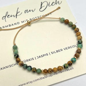 Könnte beinhalten: Verstellbares Armband mit türkisen, braunen und goldenen Perlen. Das Armband ist an einem beigen Kordelband befestigt und hat einen goldenen Perlenverschluss. Die Karte hinter dem Armband hat deutschen Text.
