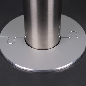 Könnte beinhalten: Nahaufnahme einer silbernen Metallbasis mit einem Puzzle-ähnlichen Design. Die Basis ist so konzipiert, dass sie um eine Metallstange passt.