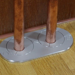 Può includere: Una piastra per pavimenti in metallo argentato con due tubi di rame che la attraversano. La piastra ha un'apertura circolare per ogni tubo e il testo "15" è inciso sulla piastra.
