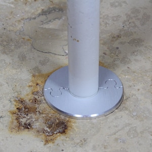 Puede incluir: Un tubo de metal blanco con una placa base plateada. La placa base tiene un diseño decorativo con recortes. El tubo está montado en un suelo de hormigón.