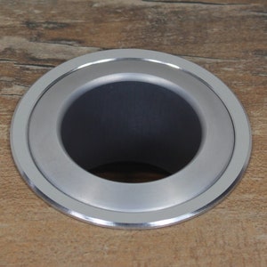 Pode incluir: Close-up de um anel de metal prateado com uma abertura circular. O anel está montado em uma superfície de madeira.