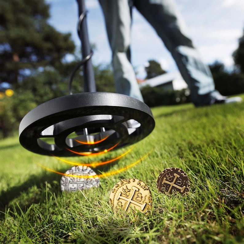 May include: A metal detector with a black handle and a large round coil is scanning the ground. Three gold coins with a cross design are visible in the grass.