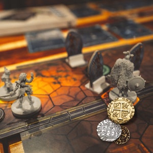 May include: A close-up of a game board with three gold and silver coins. The coins have a cross design and are on a brown and orange game board with a gray miniature figure.