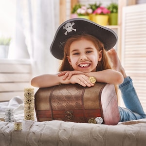 May include: A young girl wearing a black pirate hat with a skull and crossbones design is smiling and leaning on a wooden treasure chest. The chest is open and there are gold coins scattered around it.
