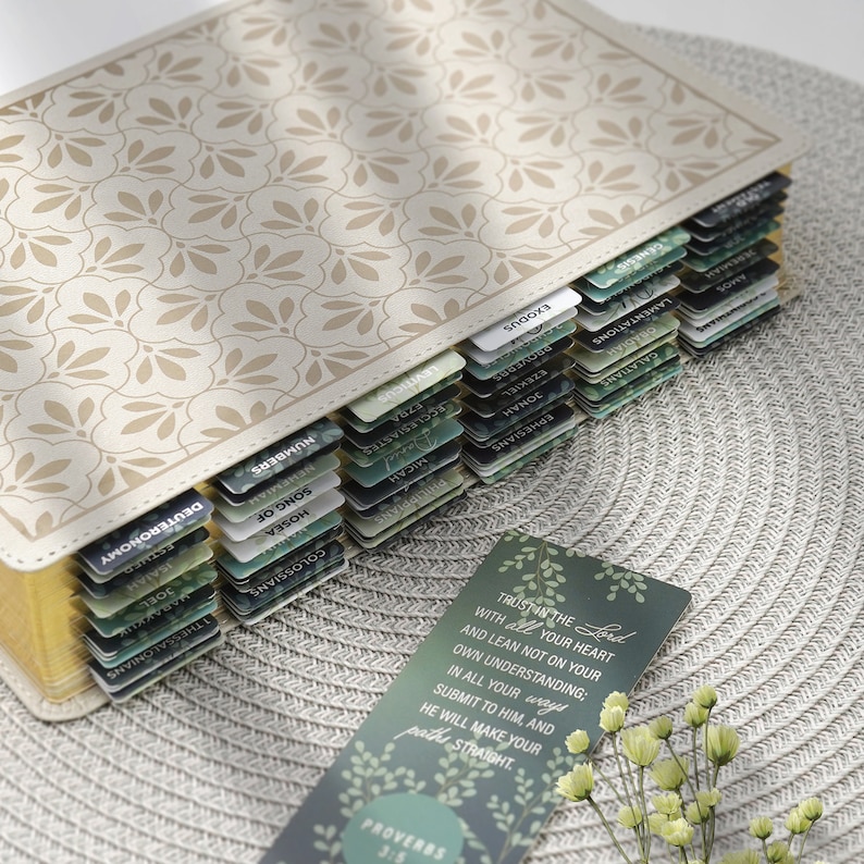 Puede incluir: Una Biblia encuadernada en cuero blanco con bordes dorados y pesta&ntilde;as verdes para cada libro de la Biblia. La Biblia est&aacute; cerrada y se encuentra sobre un mantel tejido. Un marcap&aacute;ginas verde con un dise&ntilde;o floral y el texto "Conf&iacute;a en el Se&ntilde;or con todo tu coraz&oacute;n, y no te apoyes en tu propia prudencia. Recon&oacute;celo en todos tus caminos, y &eacute;l allanar&aacute; tus sendas. Proverbios 3:5" es visible en primer plano.