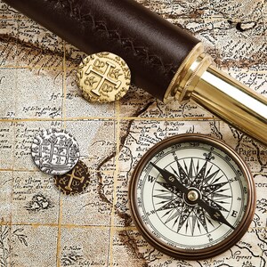 May include: A vintage compass with a black needle pointing north, surrounded by a gold-colored map with a leather-bound telescope and gold coins.
