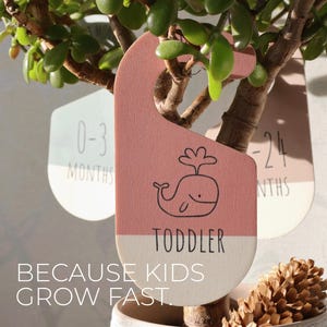 May include: A set of three wooden growth chart hangers with a whale design. The hangers are painted in pink, green, and white. The text on the hangers reads "0-3 Months", "Toddler", and "12-24 Months". The text "Because Kids Grow Fast" is printed below the hangers.