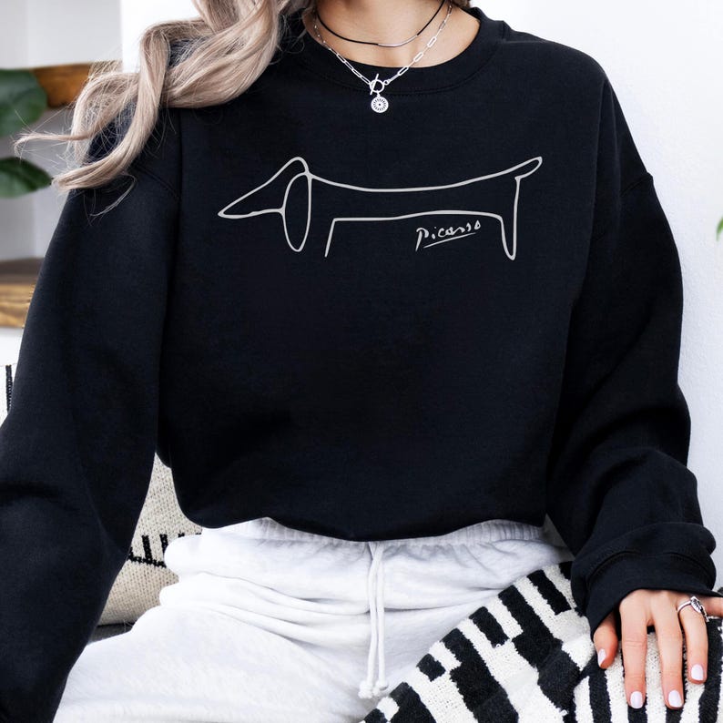May include: Black sweatshirt featuring a white line drawing of a dachshund dog in a Picasso style. The dog illustration is accompanied by the signature "Picasso" in cursive. The sweatshirt is a casual, comfortable garment.