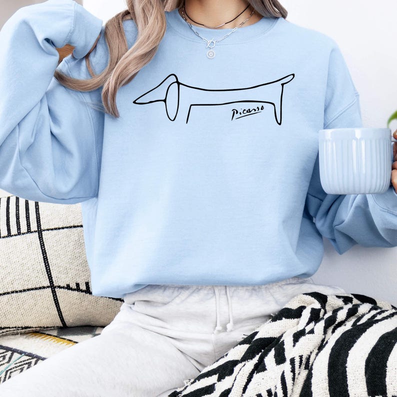 May include: Light blue sweatshirt featuring a minimalist black line drawing of a dachshund dog. The dog illustration is accompanied by the signature "Picasso" in black script. The sweatshirt is a solid light blue color.