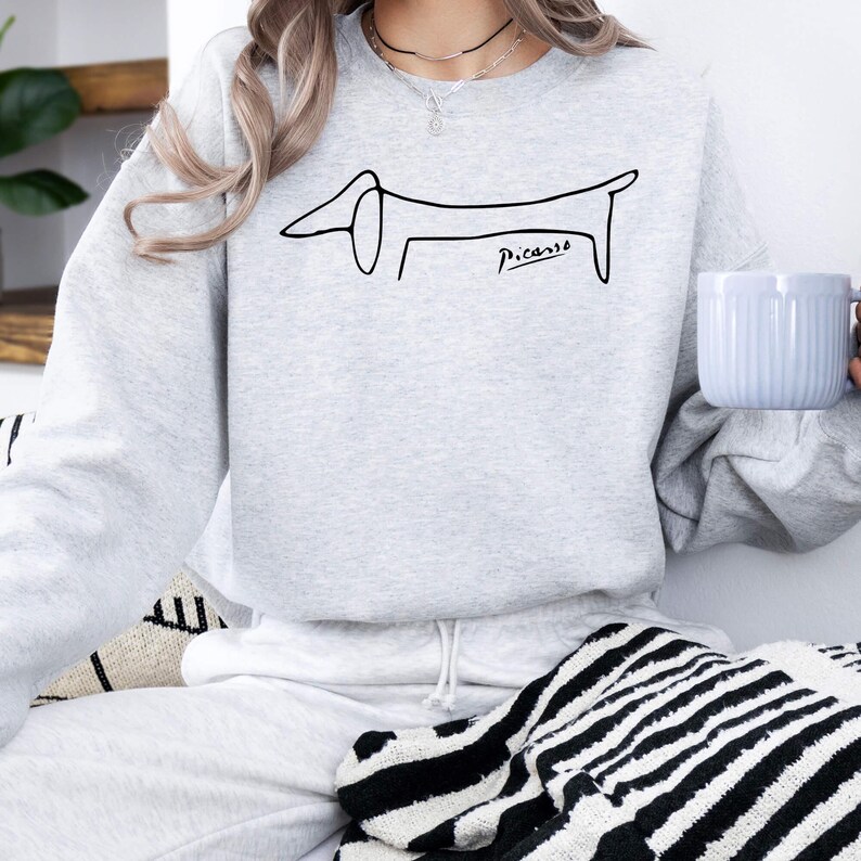 May include: A light gray sweatshirt featuring a minimalist black line drawing of a dachshund dog, with the word "Picasso" in cursive below. The sweatshirt is a comfortable, casual style.