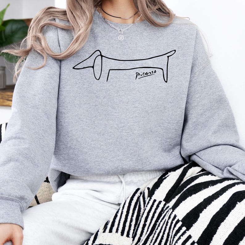 May include: A light gray sweatshirt featuring a black line drawing of a dachshund dog with the word "Picasso" in black script below the dog. The sweatshirt is a crew neck style.