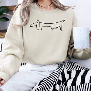 May include: Beige sweatshirt featuring a black line drawing of a dachshund dog, with the signature "Picasso" below. The sweatshirt is a neutral color, suitable for casual wear. The design is simple and artistic.