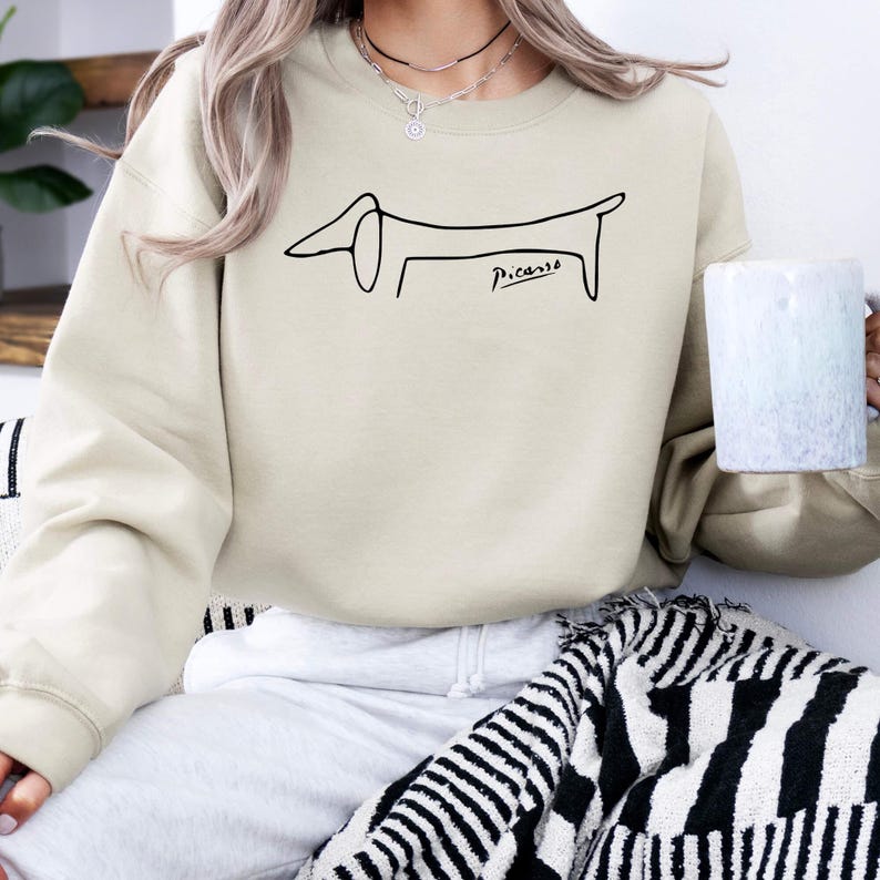 May include: Beige sweatshirt featuring a black line drawing of a dachshund dog, with the signature "Picasso" below. The sweatshirt is a neutral color, suitable for casual wear. The design is simple and artistic.