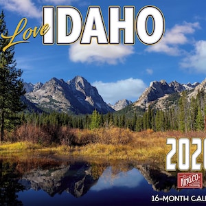 May include: A 2026 calendar featuring a scenic view of Idaho's mountains, a lake, and trees. The word "IDAHO" is displayed in large white letters with a black outline, and the words "Love" and "2026" are also visible.