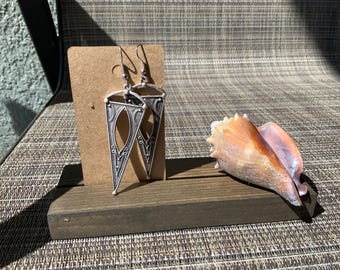 May include: A pair of silver triangle earrings with intricate designs. The earrings are displayed on a wooden stand with a seashell.
