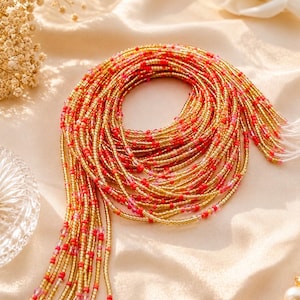 May include: A collection of multiple strands of waist beads featuring a repeating pattern of metallic gold seed beads interspersed with vibrant red and translucent pink glass beads, arranged in a coiled circular formation with loose ends.