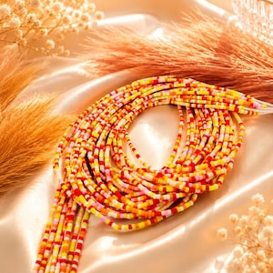 May include: A multi-strand waist bead necklace featuring small, cylindrical glass beads in a vibrant color palette of red, orange, yellow, and pink. The beads are strung on thin white threads, creating a layered, circular arrangement of colorful segments.