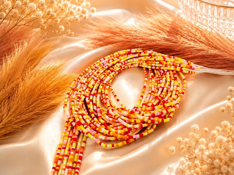 May include: A multi-strand waist bead necklace featuring small, cylindrical glass beads in a vibrant color palette of red, orange, yellow, and pink. The beads are strung on thin white threads, creating a layered, circular arrangement of colorful segments.