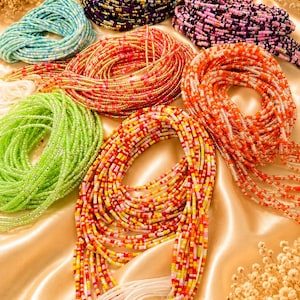 May include: A collection of multi-colored waist beads arranged in circular coils. The strands feature small glass beads in shades of light blue, dark purple, gold, orange, lime green, and a mix of red, yellow, and white, all finished with white ties.
