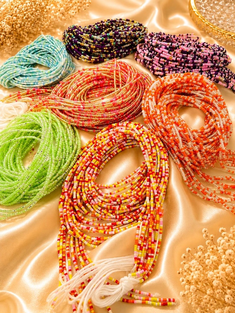 May include: A collection of multi-colored waist beads arranged in circular coils. The strands feature small glass beads in shades of light blue, dark purple, gold, orange, lime green, and a mix of red, yellow, and white, all finished with white ties.
