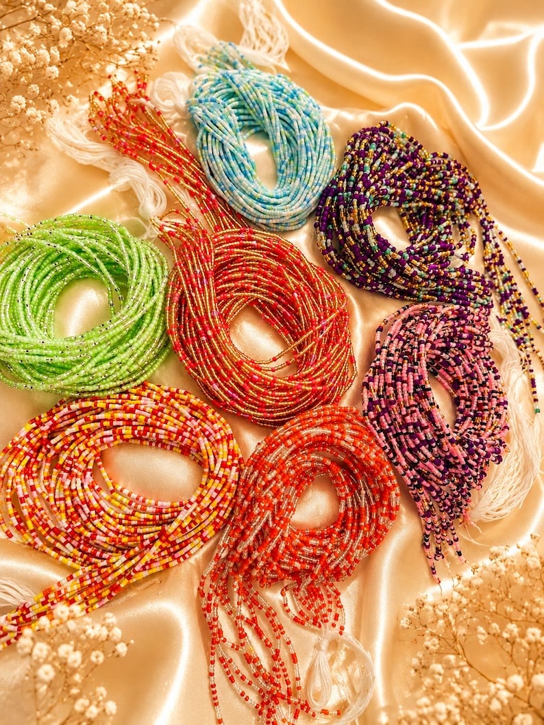 May include: A collection of seven coiled waist bead strands in various colors including light green, light blue, purple, pink, red, and multi-colored patterns. Each strand consists of small glass seed beads on a white string with loose ends.