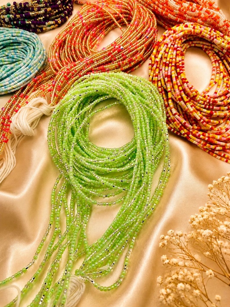 May include: A collection of waist beads featuring multiple strands of small glass beads in various colors including lime green, orange, yellow, pink, and blue, accented with metallic silver spacers and finished with white cotton tie strings.