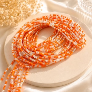 May include: A long strand of waist beads featuring a repeating pattern of opaque orange and translucent clear glass seed beads. The beads are strung on a flexible cord and coiled into a circular shape, with several loose ends hanging down.
