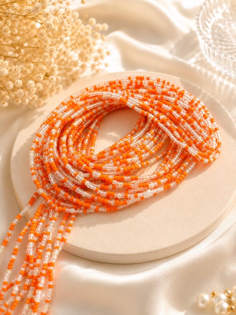 May include: A long strand of waist beads featuring a repeating pattern of opaque orange and translucent clear glass seed beads. The beads are strung on a flexible cord and coiled into a circular shape, with several loose ends hanging down.