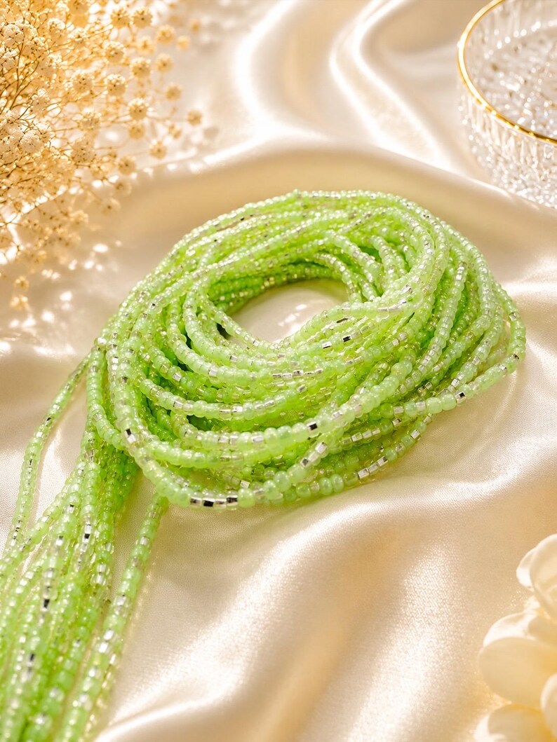 May include: A coiled strand of waist beads featuring translucent light green glass beads interspersed with small, reflective silver-colored metallic spacers. The beads are arranged in a continuous circular pattern, creating a vibrant and textured appearance.