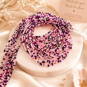 May include: A multi-strand waist bead necklace featuring small glass beads in shades of pink, purple, black, and metallic gold. A rectangular card is present with the text BEADBUTIK, Made for you. Made to shine., and HANDMADE WAISTBEADS THAT EMPOWER & ELEVATE.