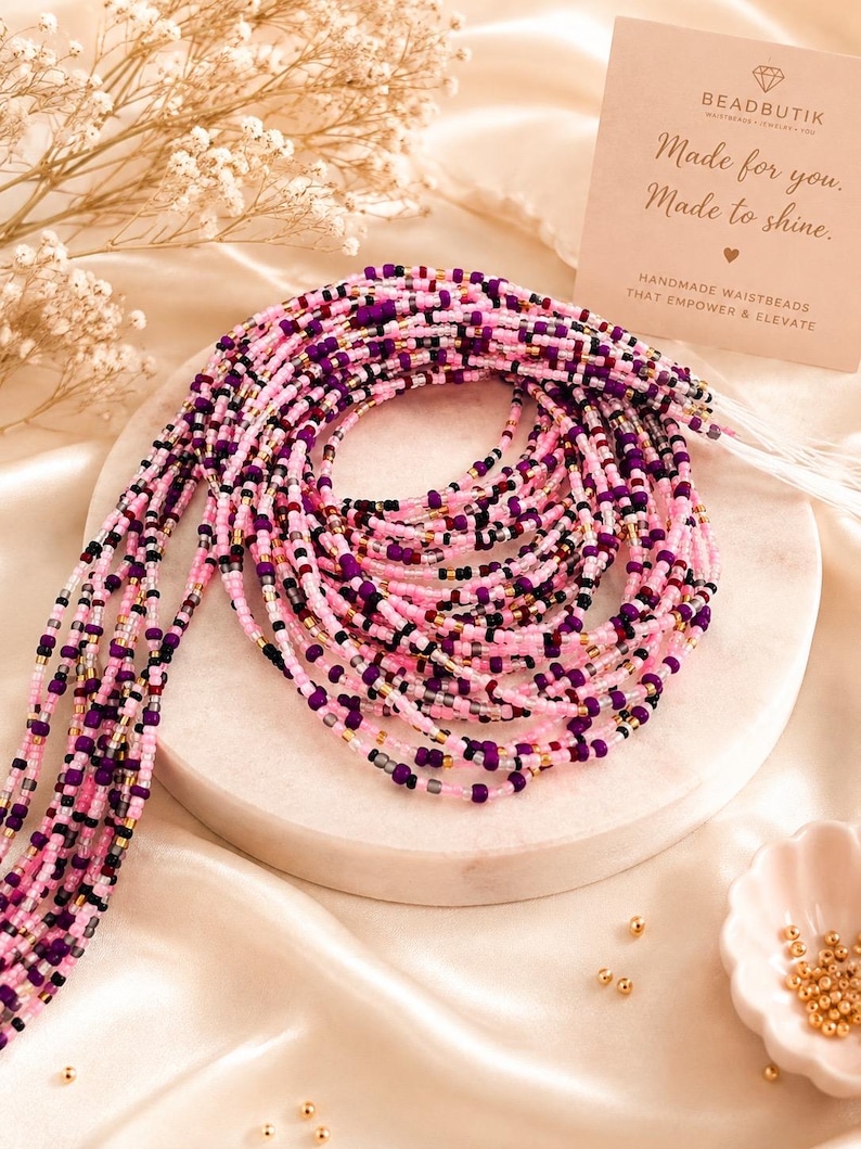 May include: A multi-strand waist bead necklace featuring small glass beads in shades of pink, purple, black, and metallic gold. A rectangular card is present with the text BEADBUTIK, Made for you. Made to shine., and HANDMADE WAISTBEADS THAT EMPOWER & ELEVATE.