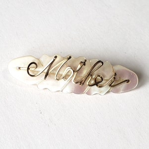 Peut inclure: Une broche en nacre blanche avec le mot "Mère" écrit en fil d'or.