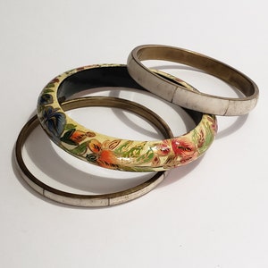 May include: Three bangle bracelets, one with a floral design painted on a cream background, one with a white bone-like material, and one with a brass finish.